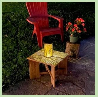 The photo depicts a small cedar table holding a solar lamp. Also in the photo are a section of a log being used as a stand for a potted plant, and a red Adirondack chair. They are on a stone patio with a portion of the lawn in the photo.