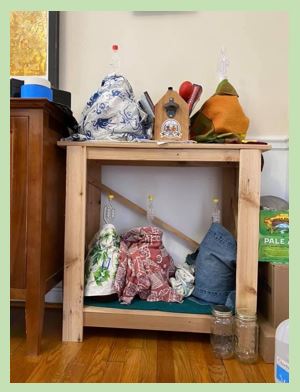 The photo depicts a small cedar table in a dining room.  The table is a rack, holding one-gallon jugs being used to make mead and cider. The jugs are covered with cloth to keep light out.