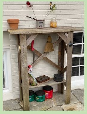 The photo depicts a chest-high table made from cedar boards. Under the table are hanging a whisk broom and a trowel for gardening and some other items.