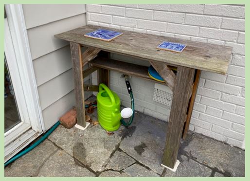 The photo shows a cedar table about waist high. It is in the corner near a sliding glass door.  It is long and narrow, with some frisbee discs and a watering can underneath.